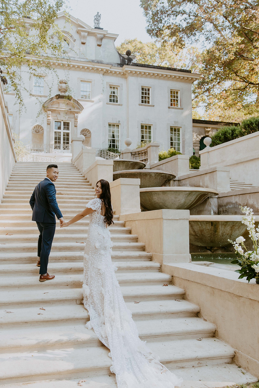 A couple stands on the steps of the Swan House, holding hands and looking back toward the camera. The woman is wearing a long white dress with a train, while the man is in a dark suit. -must have wedding poses