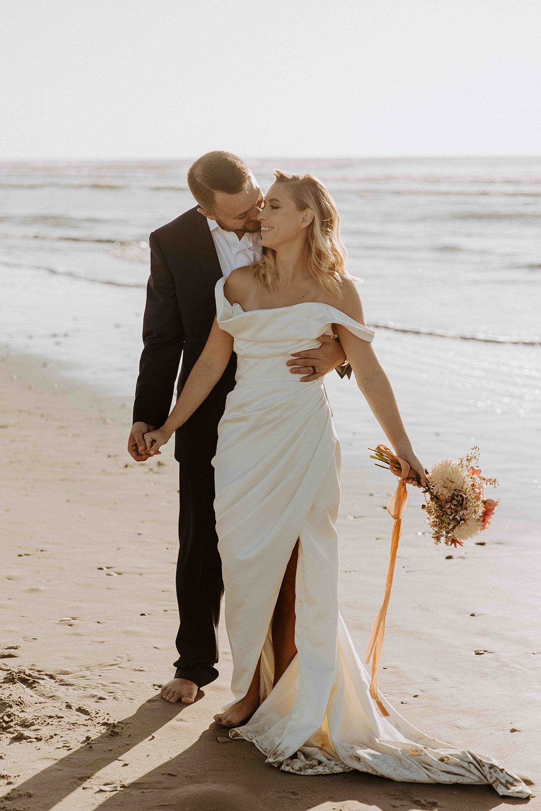 A bride and groom stand on a beach. The bride holds a bouquet and wears a white dress. The groom, in a suit, embraces her from behind as they walk barefoot on the sand near the water - must have wedding poses 