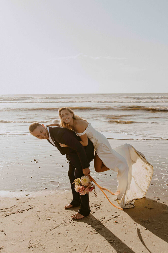 A couple in wedding attire playfully pose on a beach, with the bride on the groom's back and holding a bouquet. The groom stands on the sandy shore with the ocean in the background.