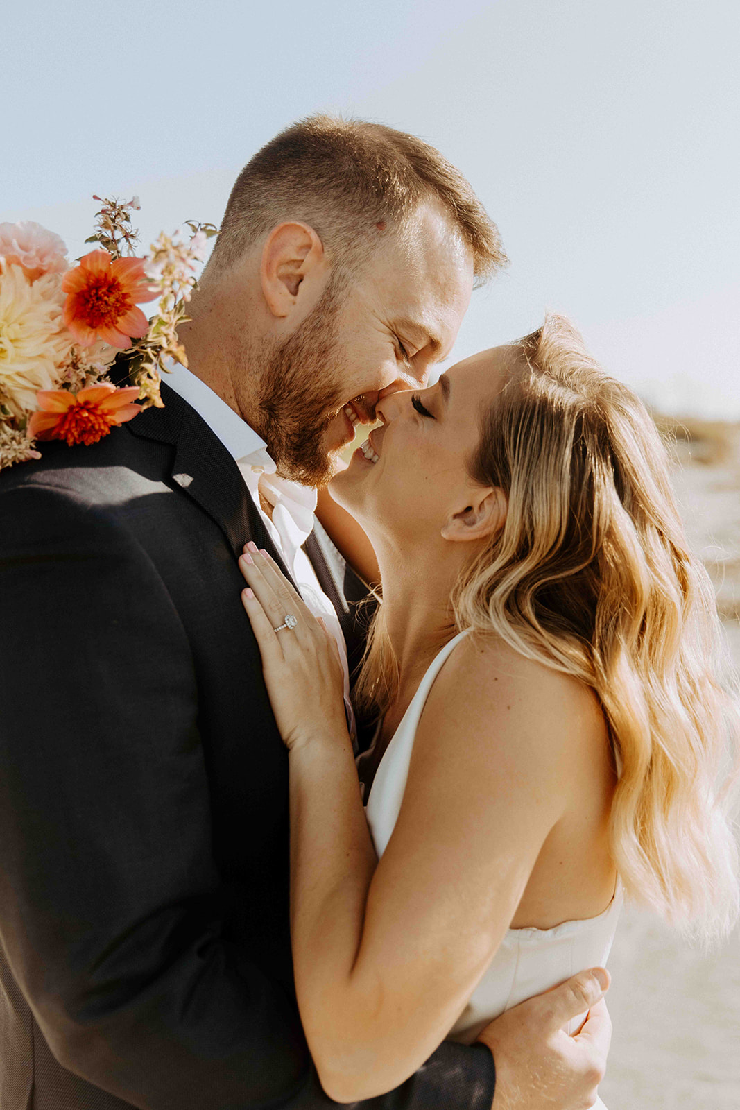 A couple in formal attire shares a kiss outdoors, with the woman holding a bouquet of flowers over the man's shoulder - must have wedding poses 