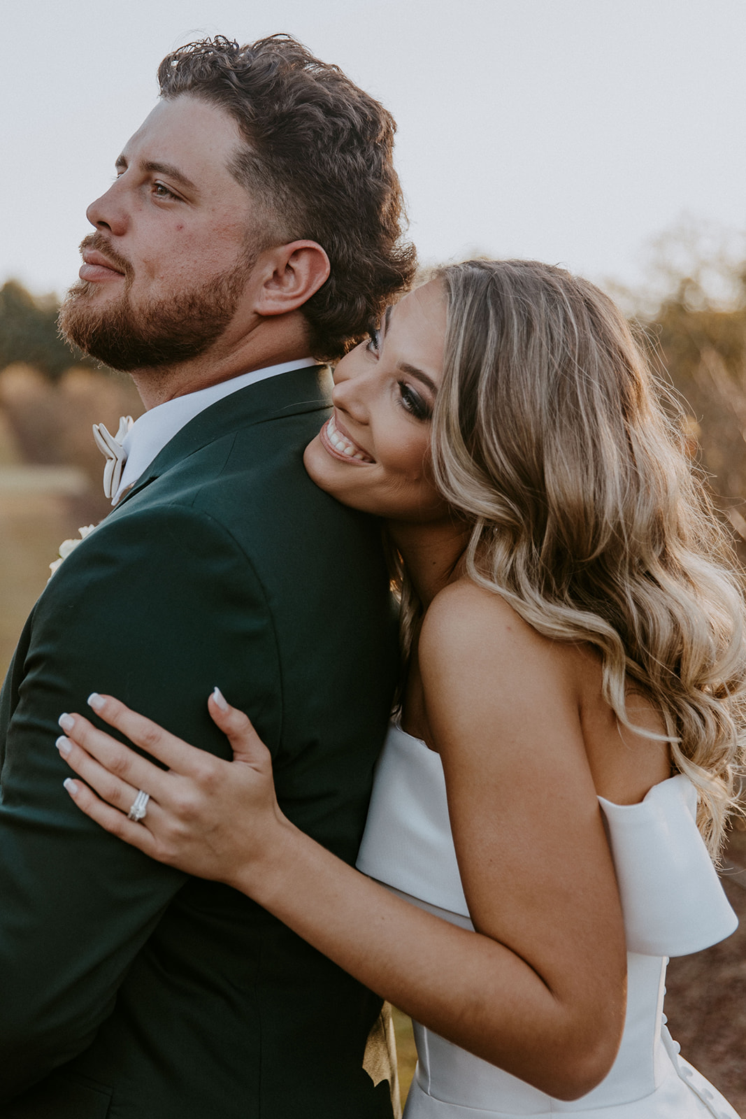 A woman in a white dress embraces a man in a green suit from behind outdoors, both smiling.