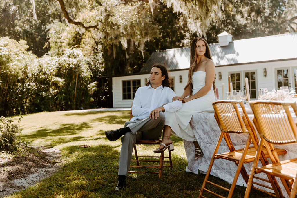 A man and a woman sit by an outdoor table with wooden chairs in a grassy yard, with a white house and trees in the background.