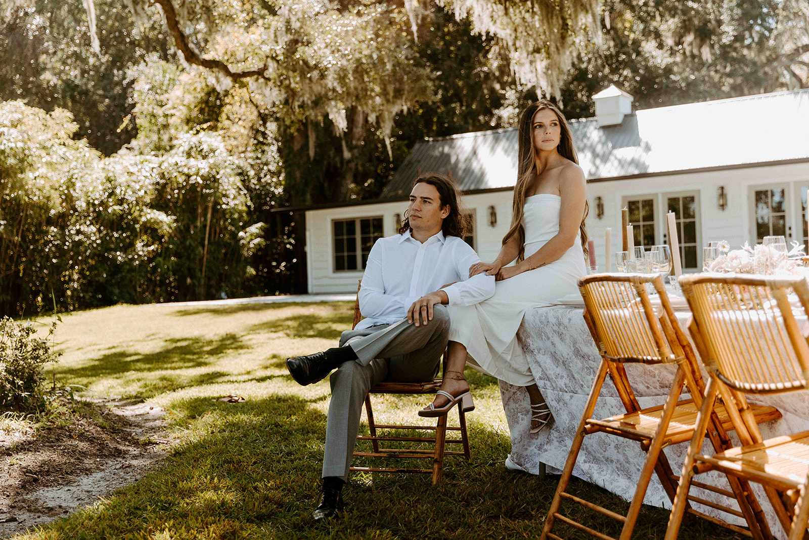 A man and a woman sit by an outdoor table with wooden chairs in a grassy yard, with a white house and trees in the background.