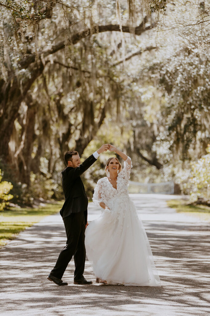 A bride in a white gown and a groom in a dark suit hold hands and dance outdoors on a grassy path, surrounded by autumnal trees. -must have wedding poses