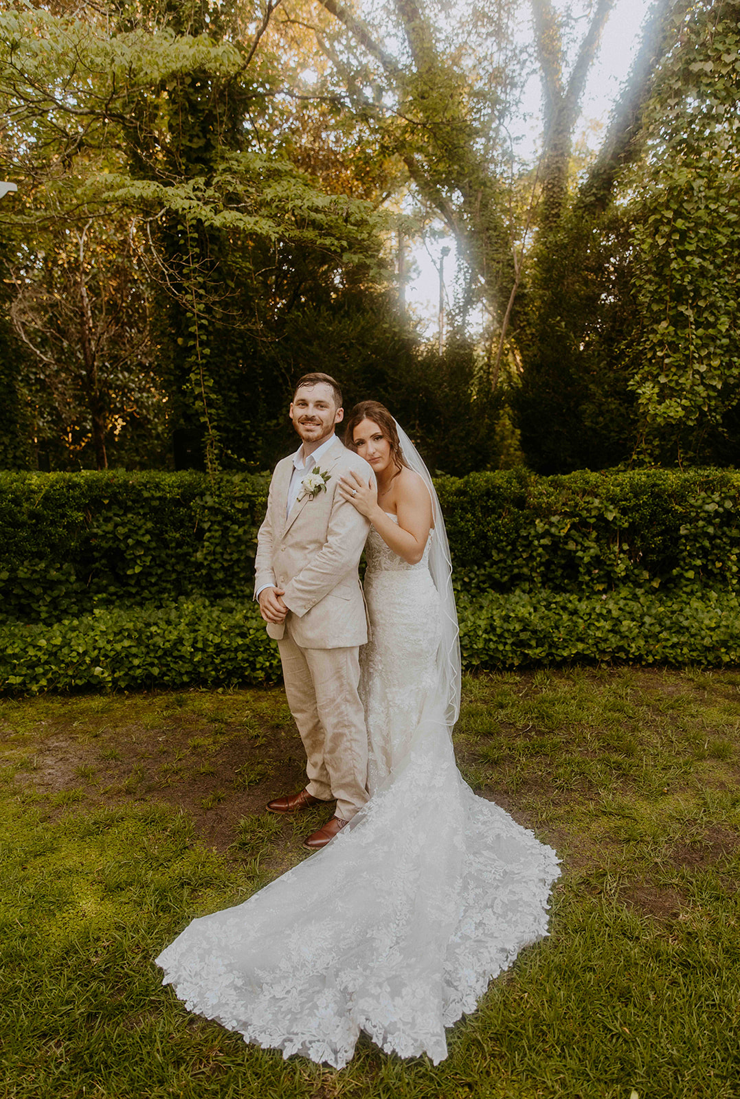 A bride and groom stand together outdoors in a garden, with the bride in a long white gown and veil holding the groom's shoulders, and the groom wearing a beige suit.