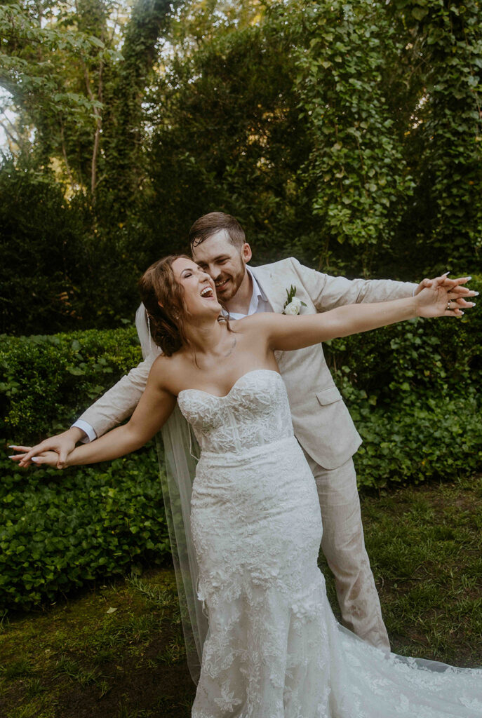 A couple in wedding attire poses outdoors. The woman, in a white strapless gown, extends her arms, while the man, in a light-colored suit, stands behind her, also with arms outstretched.