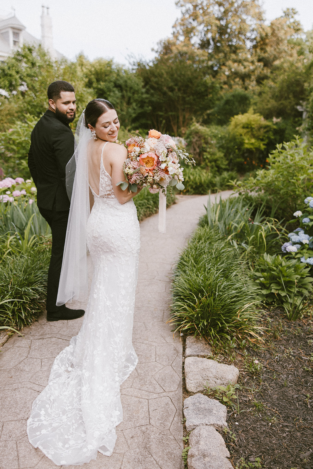 A bride in a white lace gown and veil holds a bouquet while standing on a garden path, looking back. A groom in a black suit stands behind her -must have wedding poses