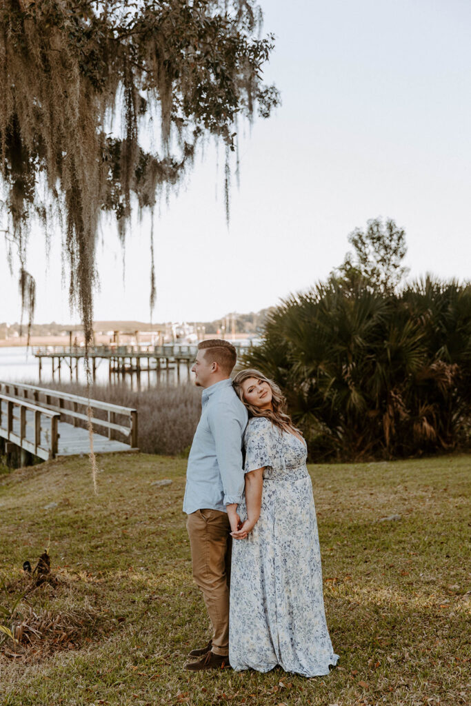 A person kneels on one knee holding an item, while another person in a floral dress stands facing them. They are outdoors near a tree with hanging moss and a body of water in the background for a surprise proposal