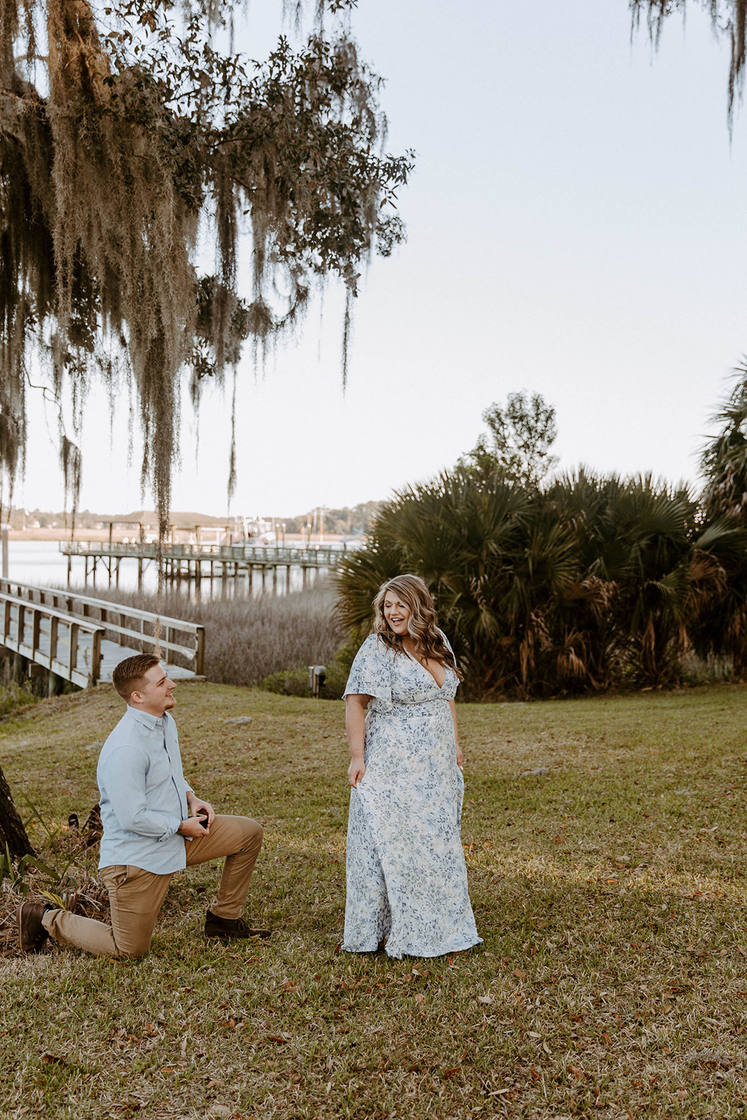 A person kneels on one knee holding an item, while another person in a floral dress stands facing them. They are outdoors near a tree with hanging moss and a body of water in the background.