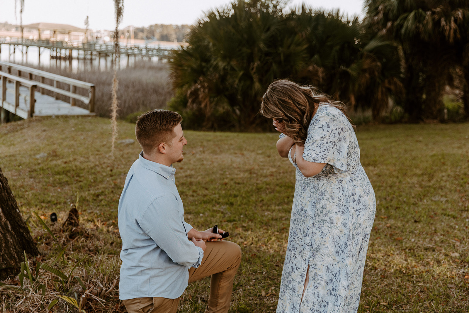 A person kneels on one knee holding an item, while another person in a floral dress stands facing them. They are outdoors near a tree with hanging moss and a body of water in the background.