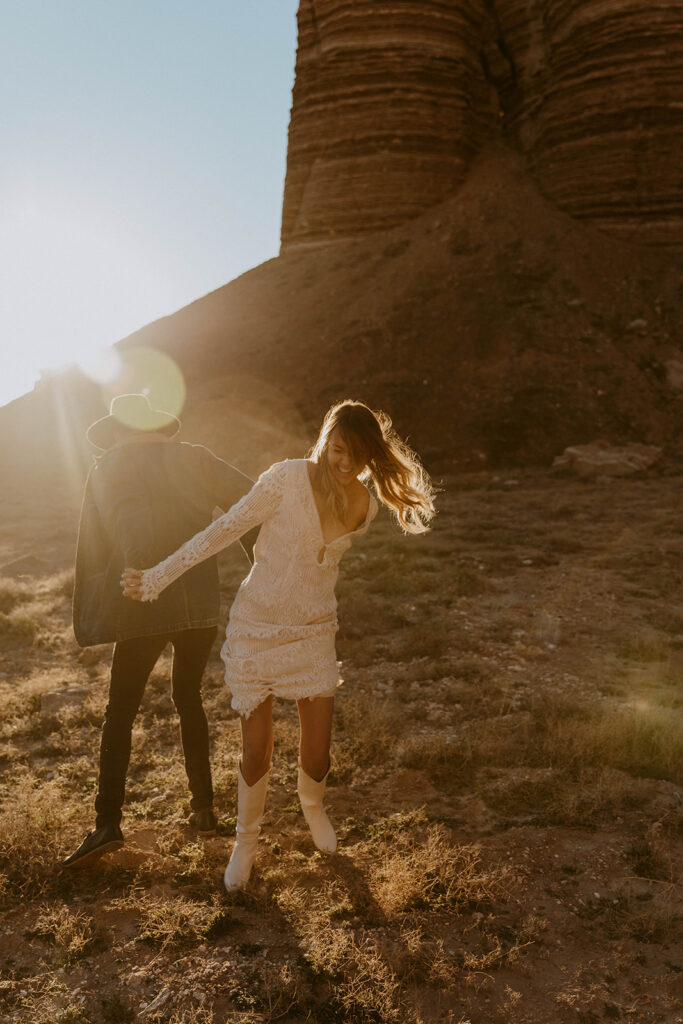 A person in a white outfit holds hands with another person in a dark outfit. They are outdoors near rocky terrain with sunlight streaming from behind.