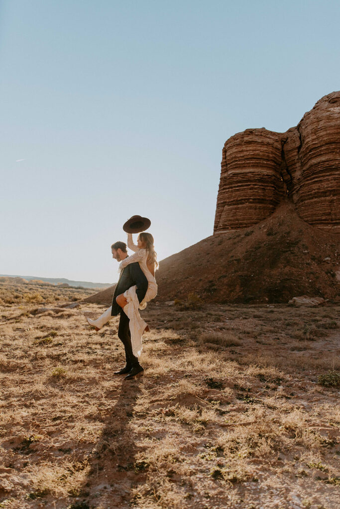 A couple is standing in a desert landscape with large rock formations in the background. The woman is on the man's back, holding a large rock over her head. Both are looking outwards.