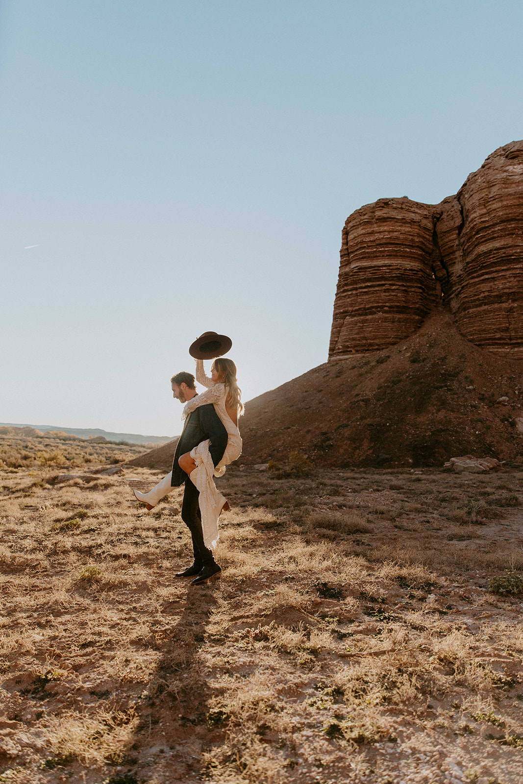 A couple is standing in a desert landscape with large rock formations in the background. The woman is on the man's back, holding a large rock over her head. Both are looking outwards The best wedding and elopement timeline examples