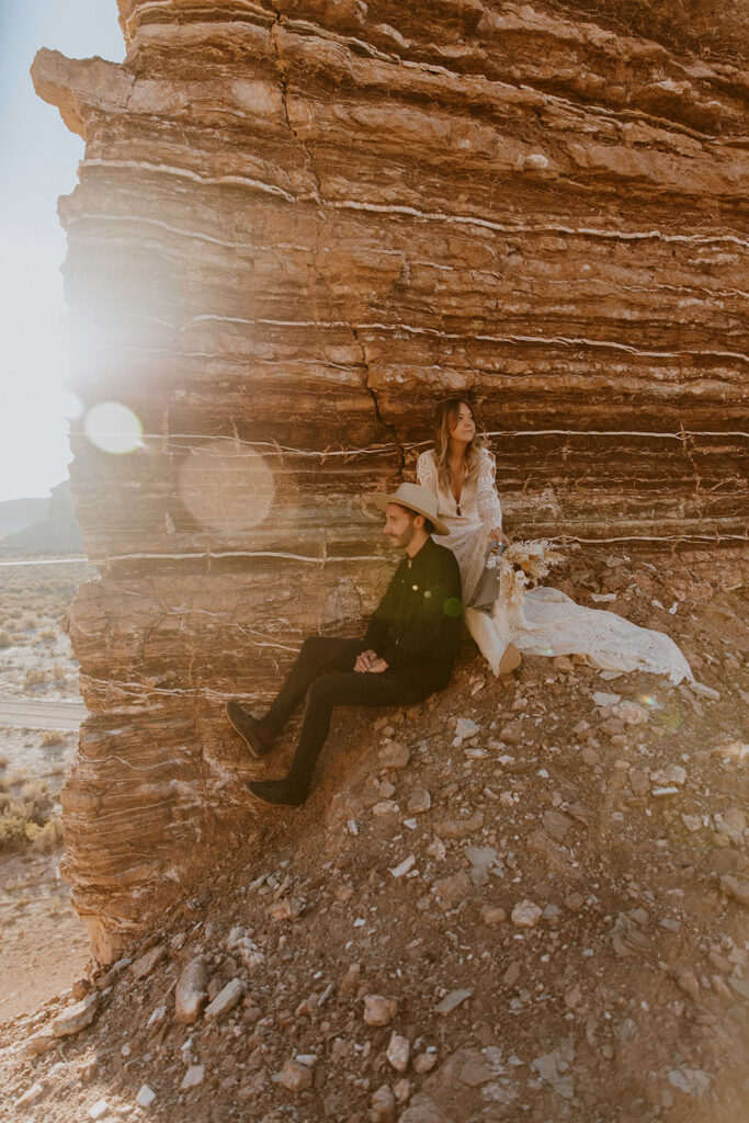 A couple, dressed in formal attire, sits together on a rocky slope beside a layered rock formation, with sunlight flaring in the background.