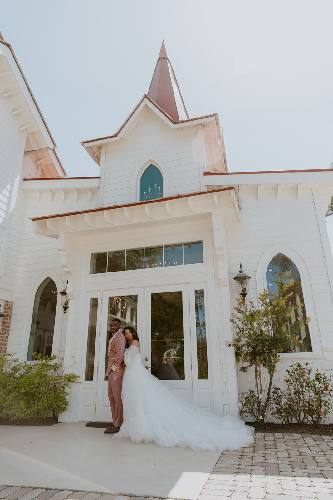 A bride and groom stand close together in front of a white, chapel-style building with arched windows and a steeple on a sunny day.