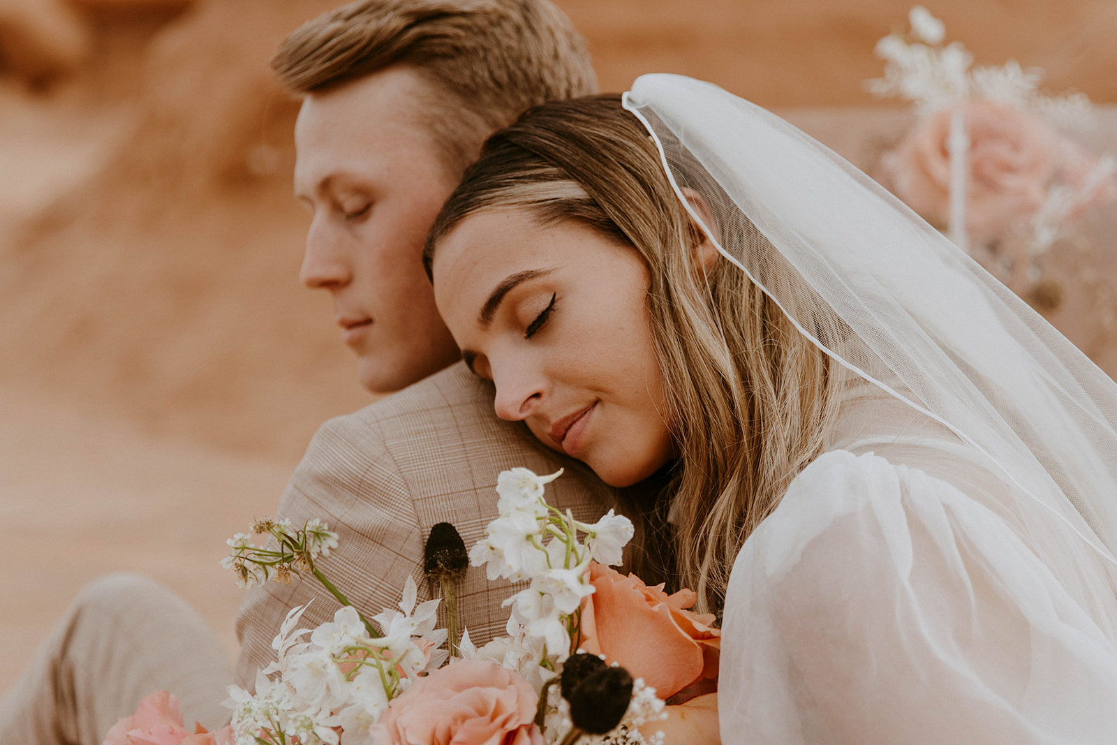 A bride in a white dress and veil rests her head on the groom's shoulder. Both are holding a bouquet of flowers.