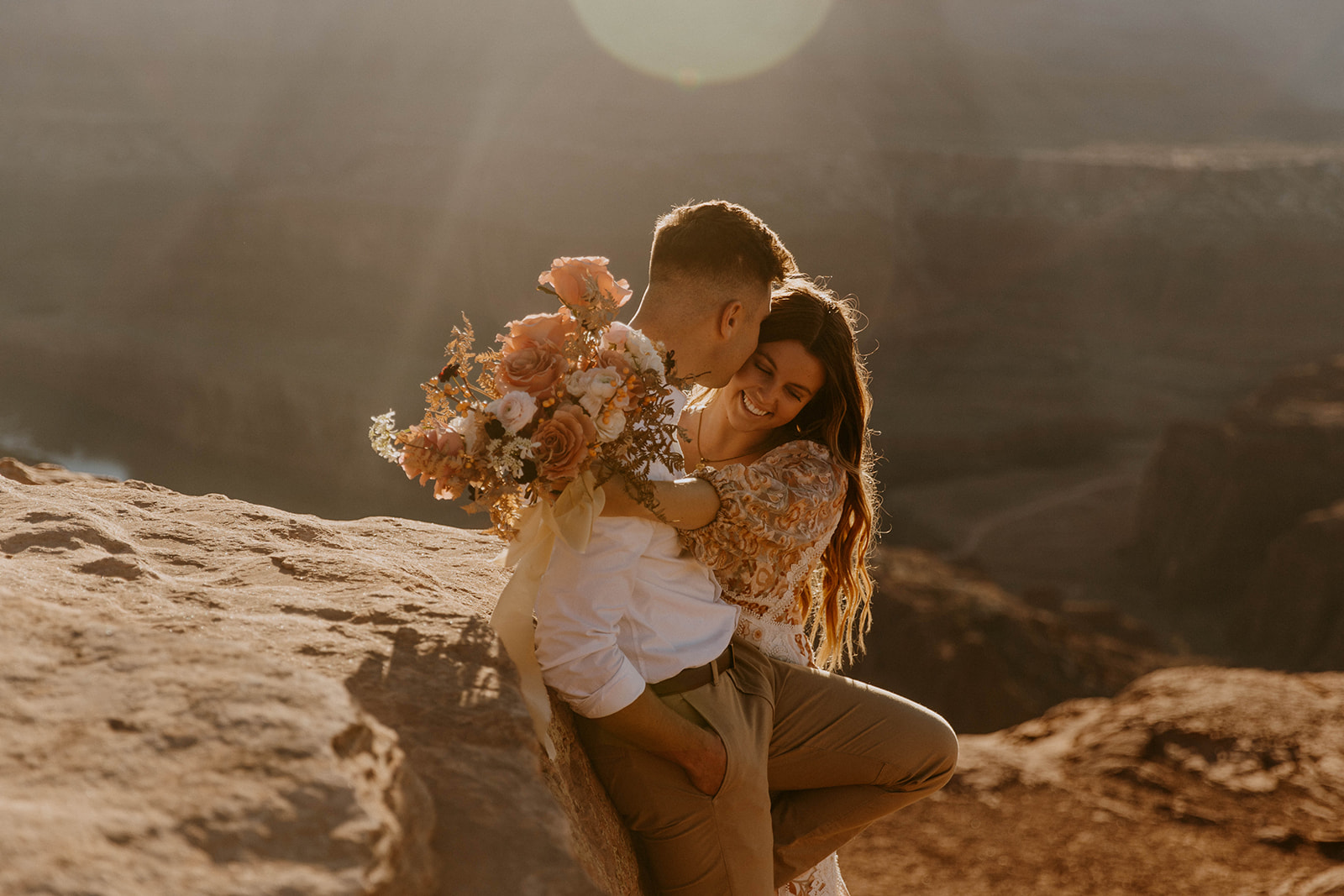 A couple sits on a rock at sunset, with the man holding a bouquet of flowers and the woman leaning against him - must have wedding poses