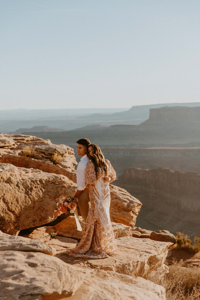 A couple dressed in casual attire stands on a rocky cliff overlooking a vast canyon landscape.