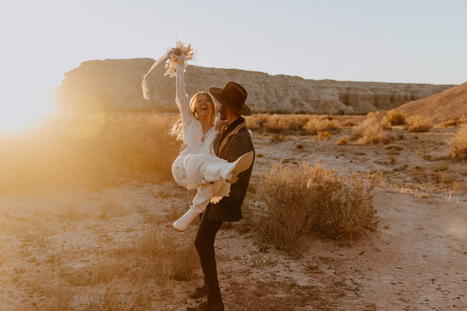 A couple in wedding attire, with the bride being lifted by the groom, pose in a sunlit desert landscape. The bride holds a bouquet aloft, and dry vegetation is scattered around them. -must have wedding poses