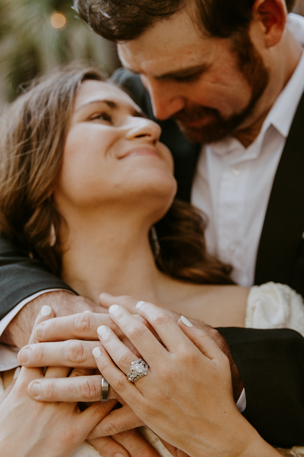 A couple stands together, the woman displaying an engagement ring on her hand as she looks up at the man who has his arms wrapped around her.