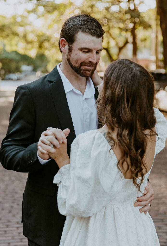 A bride in a white gown and a groom in a dark suit hold hands and dance outdoors on a grassy path, surrounded by autumnal trees. -must have wedding poses