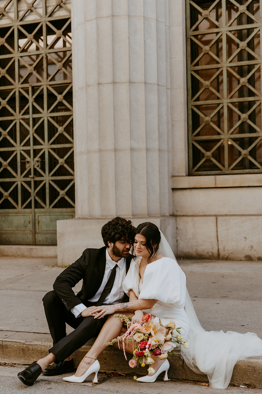 A bride and groom sit on steps outside a large building with tall columns, the groom in a black suit and the bride in a white dress holding a colorful bouquet - must have wedding poses