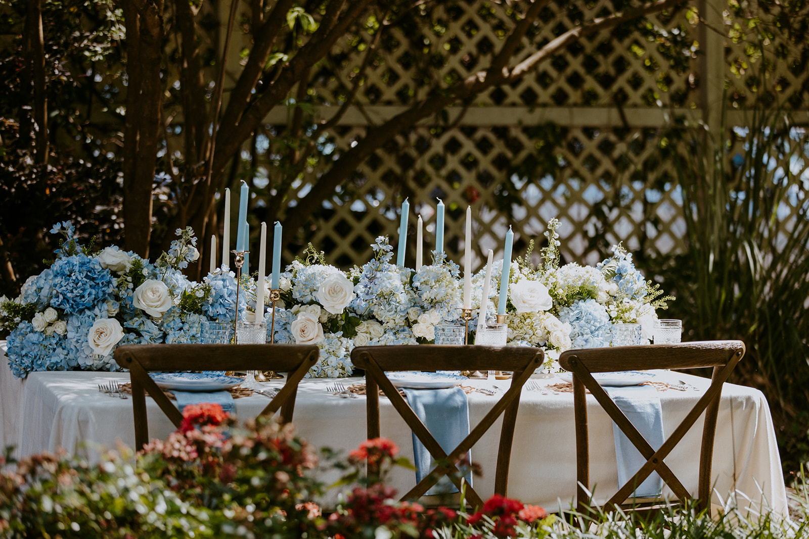 A table set for an outdoor event with wooden chairs, white tablecloth, blue accents, floral arrangements of white and blue flowers, and tall white candles in a garden setting.