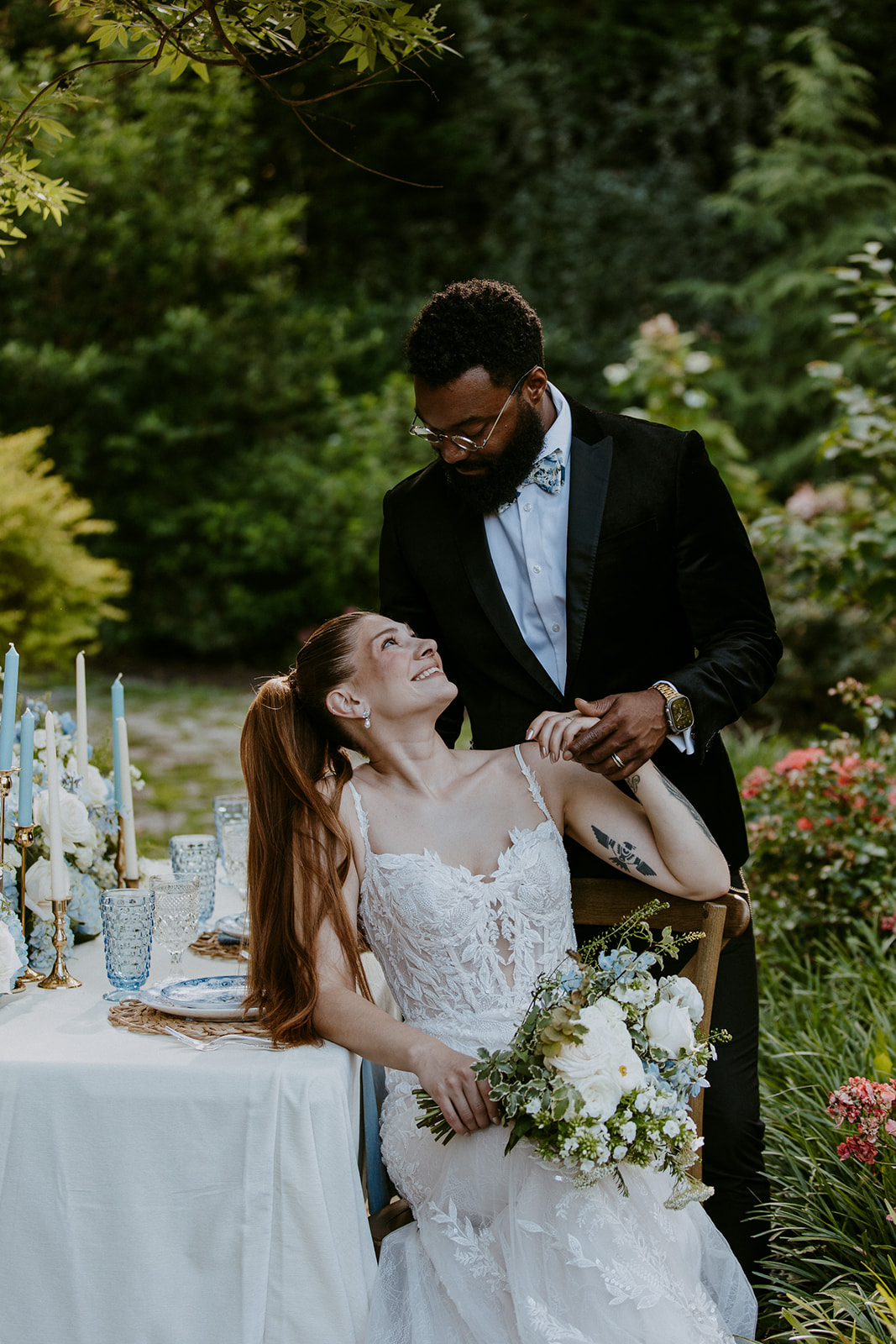 A bride and groom pose in a garden. The bride is seated with a bouquet, wearing a lace wedding dress. The groom, in a black suit, stands beside her, resting one hand on her shoulder while she looks up at him.