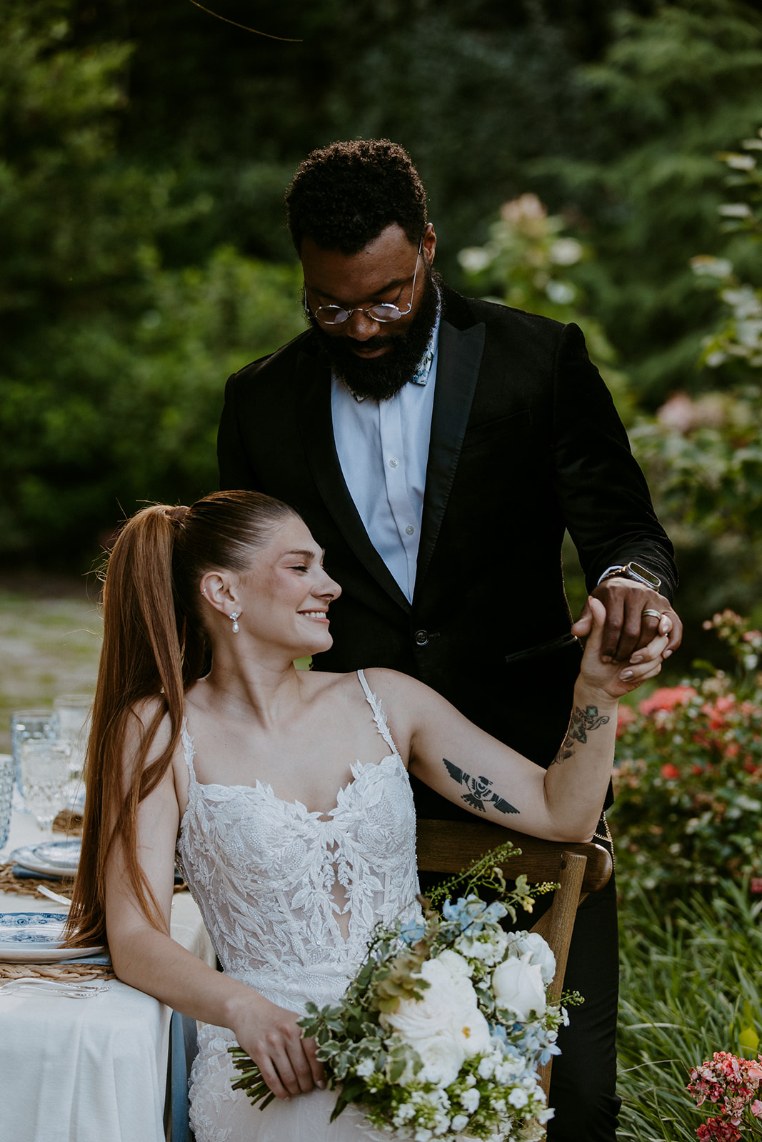 A bride and groom pose in a garden. The bride is seated with a bouquet, wearing a lace wedding dress. The groom, in a black suit, stands beside her, resting one hand on her shoulder while she looks up at him.