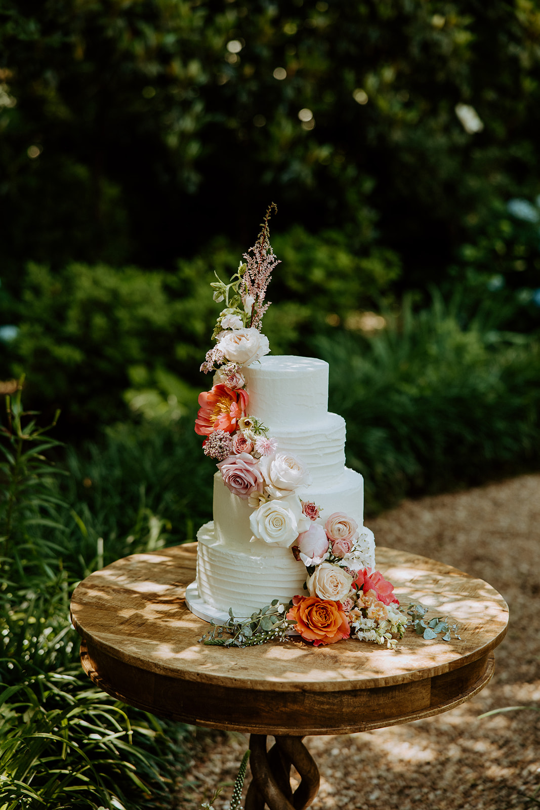 A three-tiered white wedding cake adorned with various pink, white, and orange flowers is placed on a wooden table outdoors, surrounded by greenery at Wildflower 301