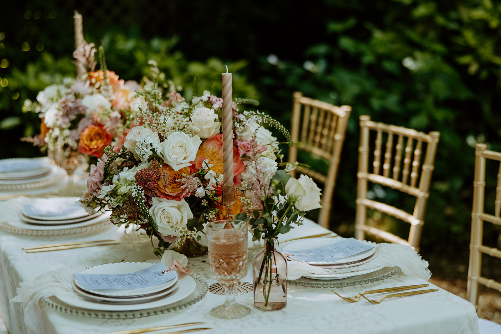 A table decorated with floral centerpieces, gold cutlery, and elegant place settings, set outdoors with three wooden chairs.