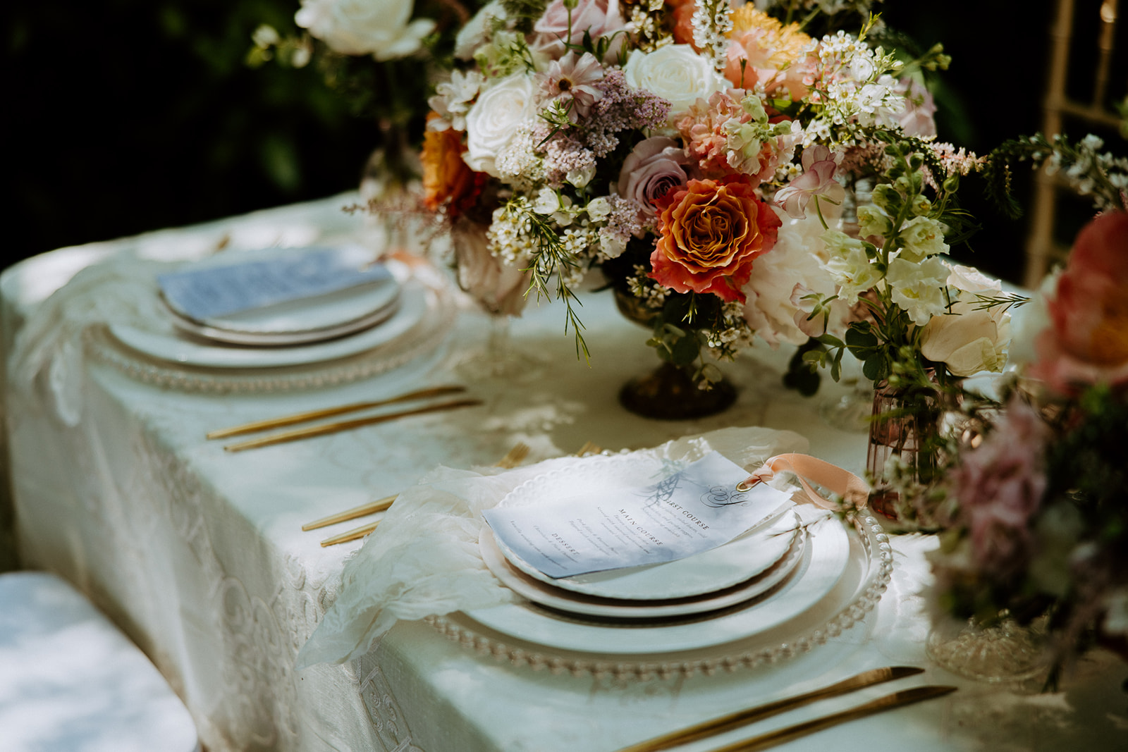 A table decorated with floral centerpieces, gold cutlery, and elegant place settings, set outdoors with three wooden chairs.