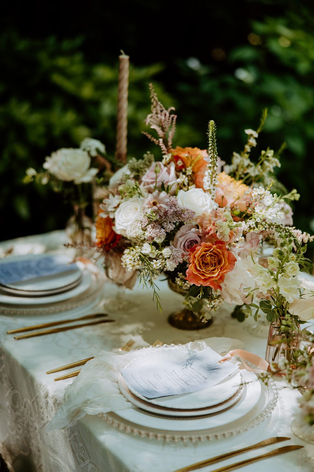 A table decorated with floral centerpieces, gold cutlery, and elegant place settings, set outdoors with three wooden chairs.