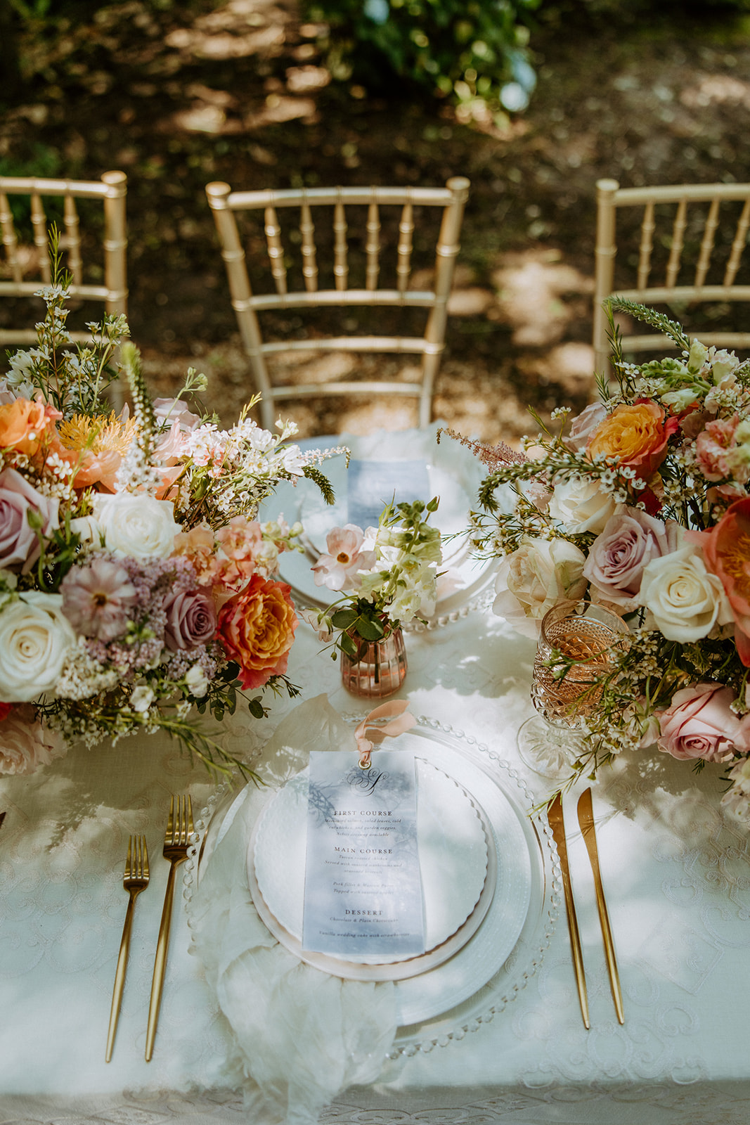 A table decorated with floral centerpieces, gold cutlery, and elegant place settings, set outdoors with three wooden chairs.