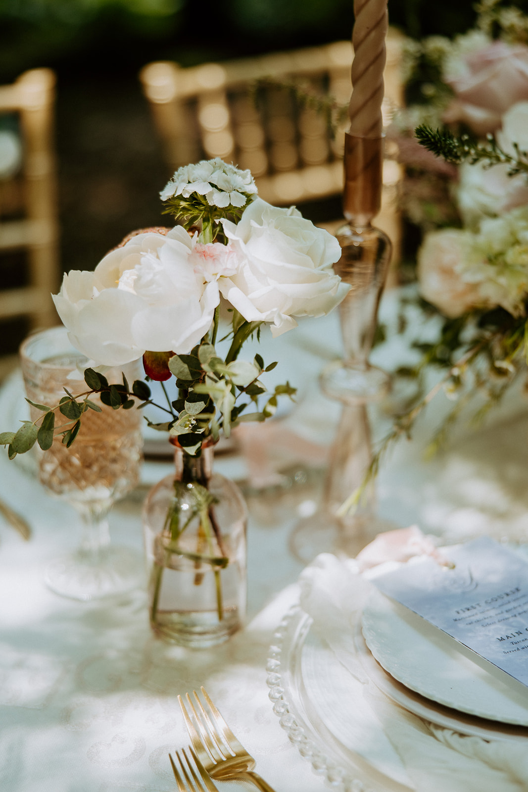 A table decorated with floral centerpieces, gold cutlery, and elegant place settings, set outdoors with three wooden chairs.