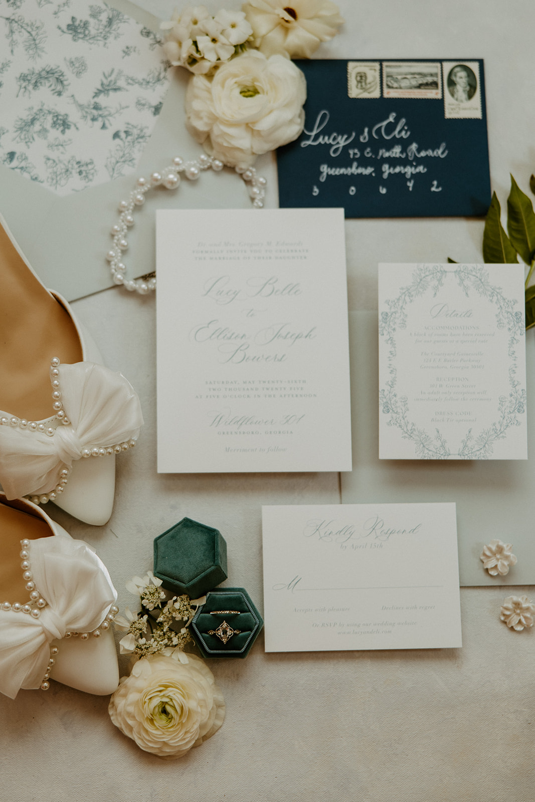 Flat lay of wedding invitations, a pair of white shoes with bows, a pearl necklace, floral accessories, a dark blue envelope, and two green ring boxes with rings, against a light background at Wildflower 301