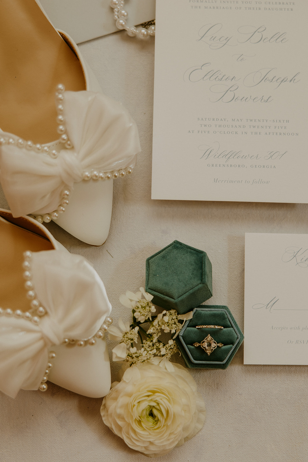 Flat lay of wedding invitations, a pair of white shoes with bows, a pearl necklace, floral accessories, a dark blue envelope, and two green ring boxes with rings, against a light background at Wildflower 301