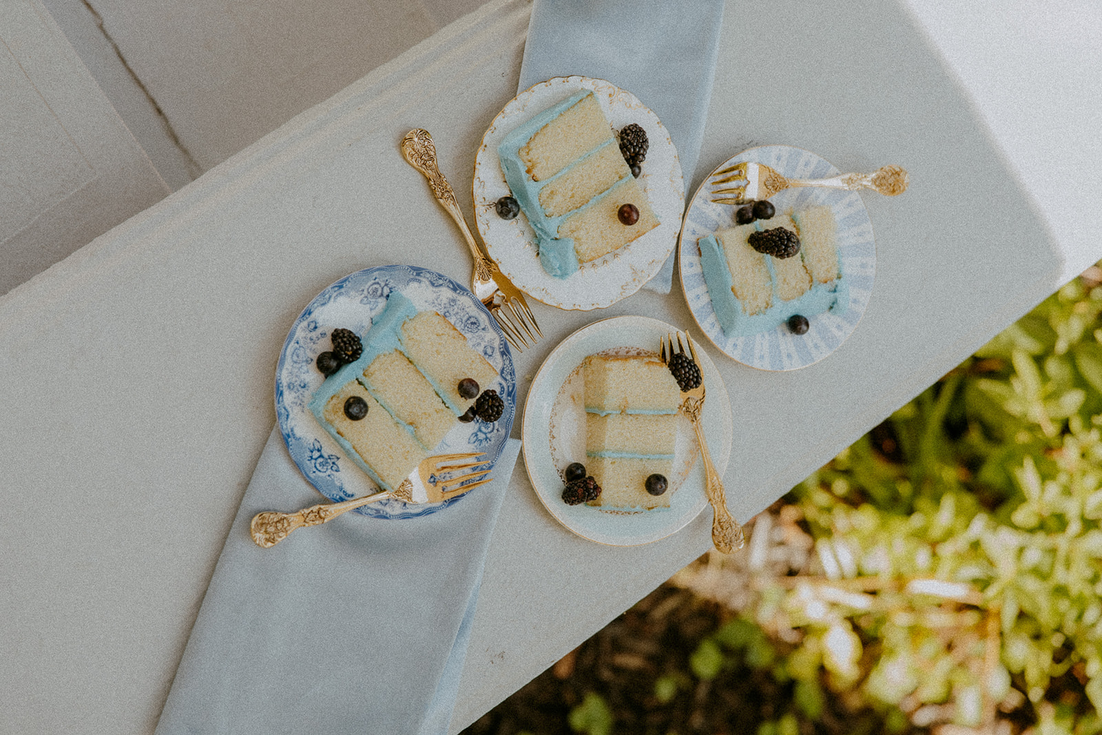 Flat lay of wedding invitations, a pair of white shoes with bows, a pearl necklace, floral accessories, a dark blue envelope, and two green ring boxes with rings, against a light background at Wildflower 301