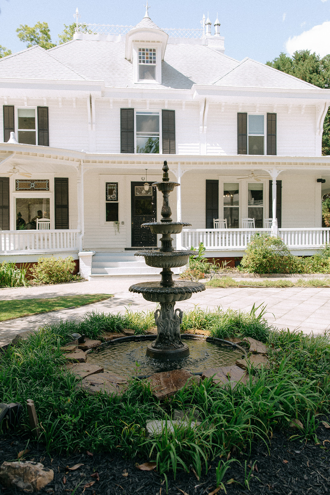 A two-story white house with black shutters and a porch is pictured behind a three-tiered stone fountain surrounded by greenery at Wildflower 301