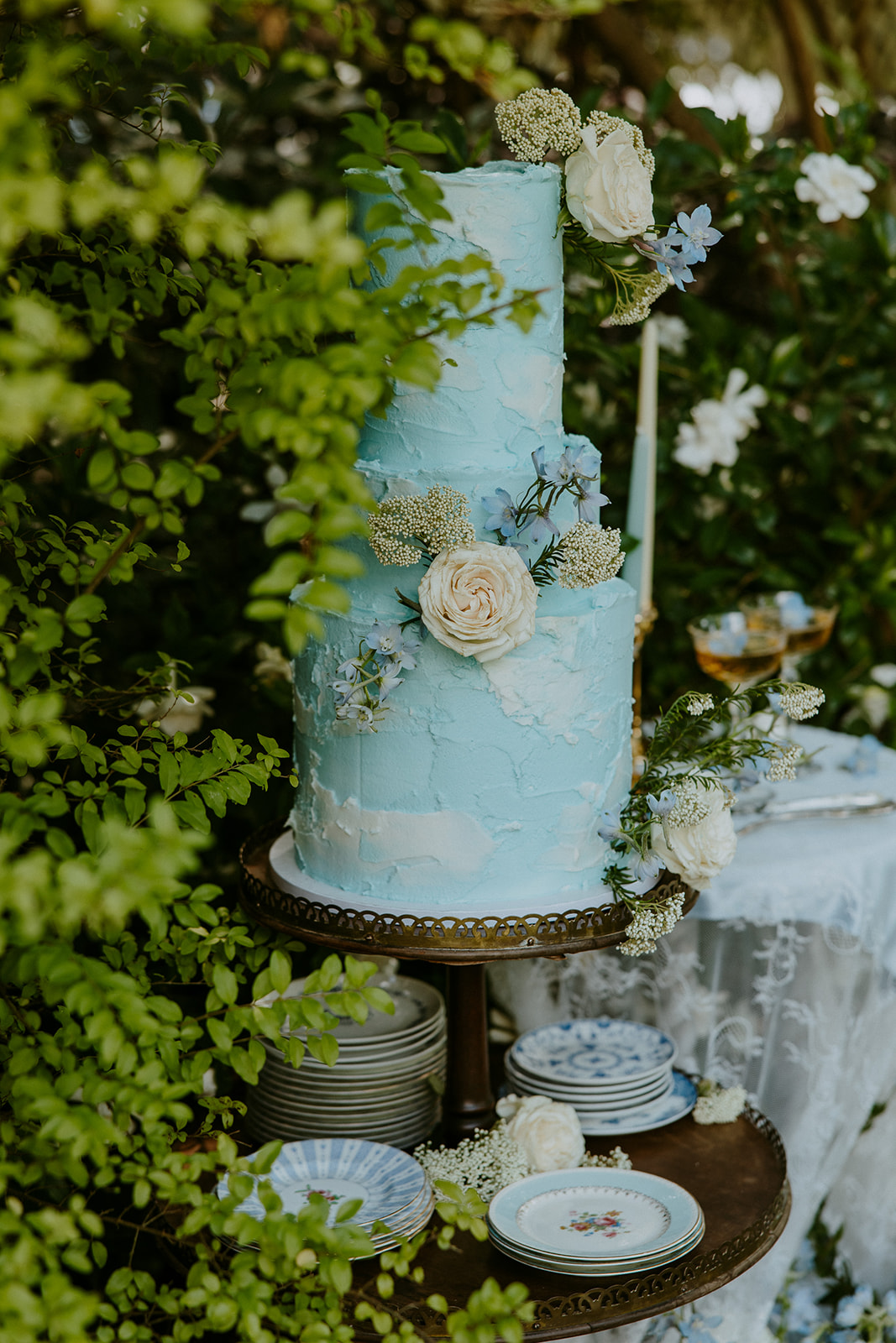 A small outdoor wedding cake table with a three-tiered cake, candles, plates, and floral decorations surrounded by blooming white flowers and greenery.