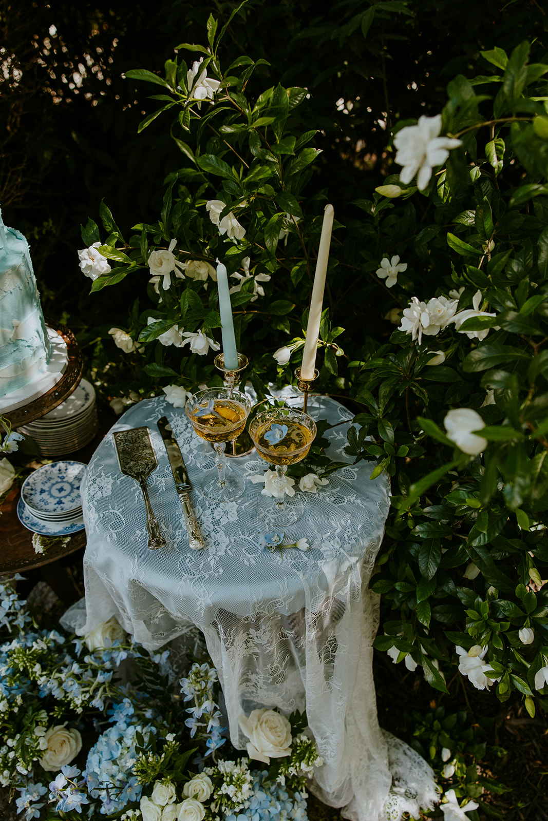 A small outdoor wedding cake table with a three-tiered cake, candles, plates, and floral decorations surrounded by blooming white flowers and greenery.