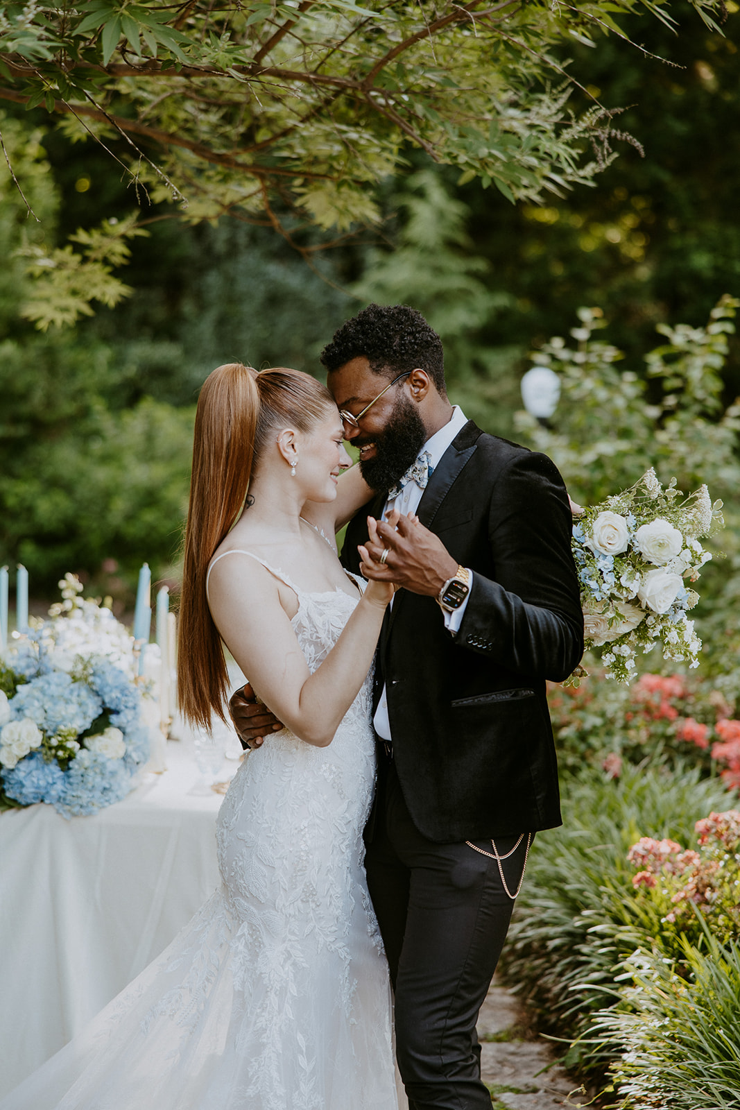 A couple stands outdoors in a garden setting, at Wildflower 301 wearing wedding attire. They are smiling and holding champagne glasses. A decorated table with a blue and white cake is beside them