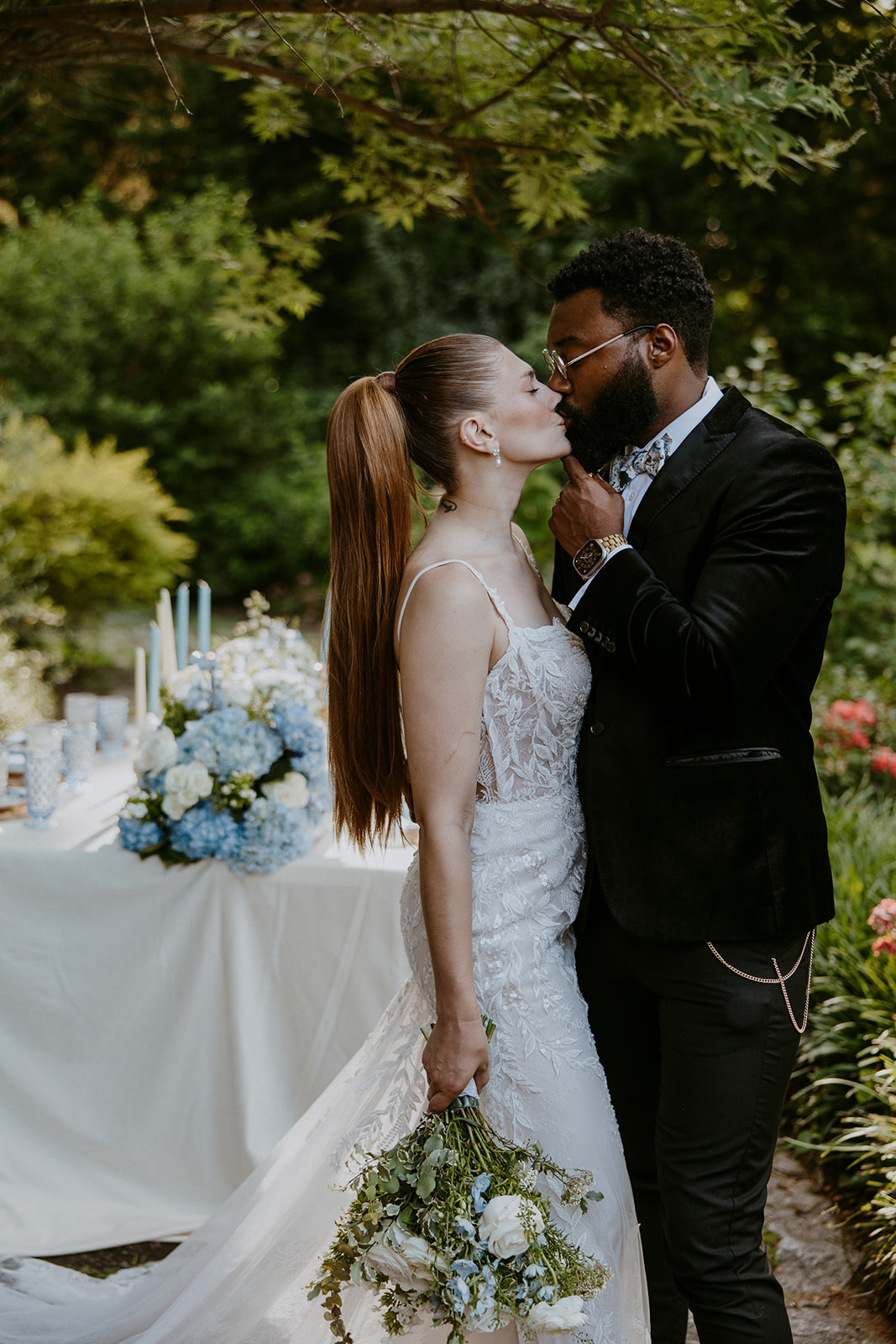 A bride and groom share a tender moment outdoors, with the bride holding a bouquet and the groom touching her chin. A table with floral decorations and candles is in the background.