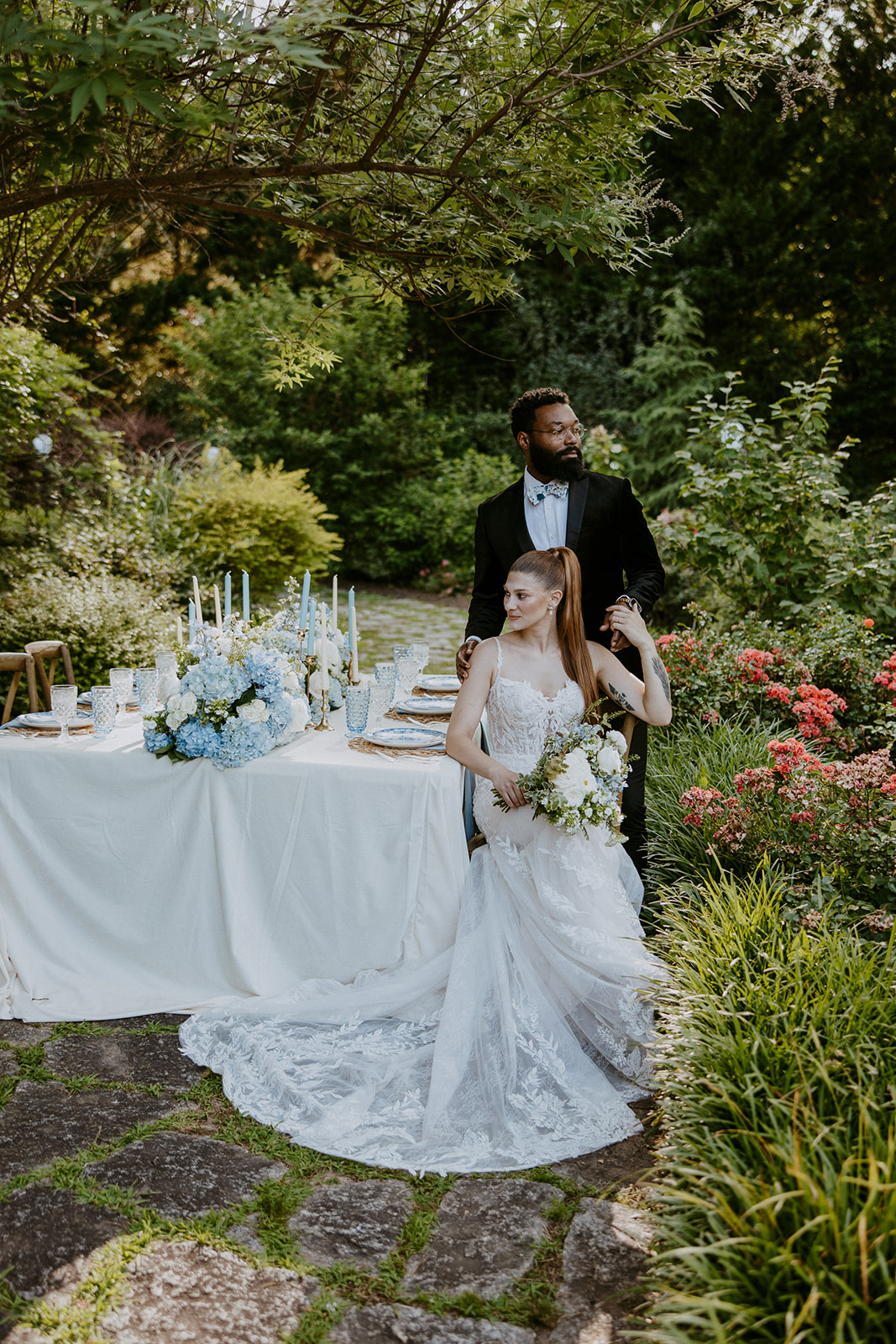 A bride and groom share a tender moment outdoors, with the bride holding a bouquet and the groom touching her chin. A table with floral decorations and candles is in the background.