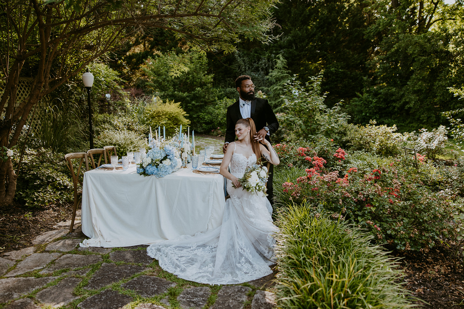 A bride in a white gown sits holding flowers while a groom in a black tuxedo stands behind her, both positioned beside an elegantly set table outdoors with foliage and flowers around.