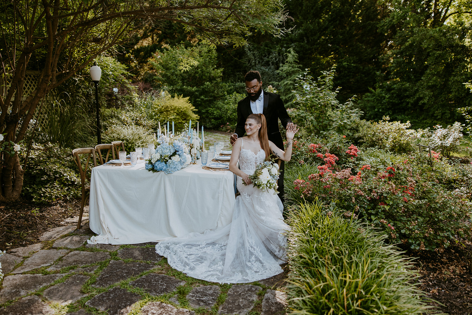 A bride in a white gown sits holding flowers while a groom in a black tuxedo stands behind her, both positioned beside an elegantly set table outdoors with foliage and flowers around. | The best wedding and elopement timeline examples