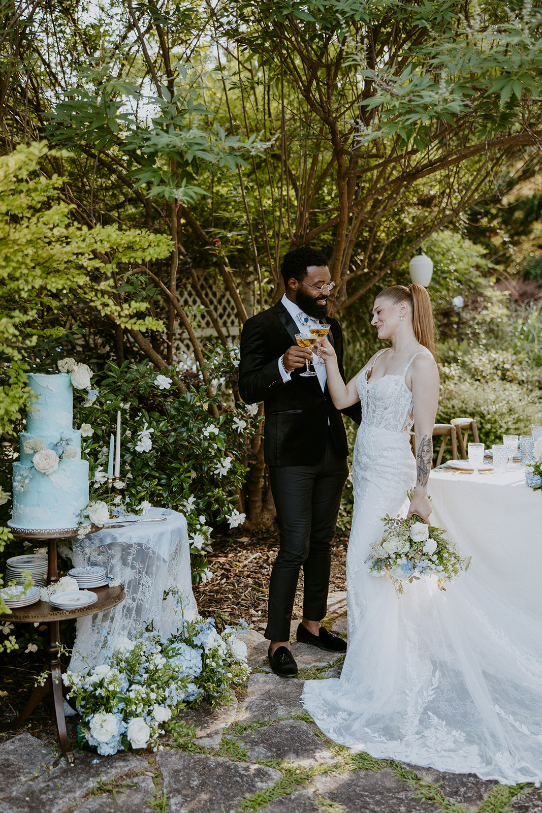 A couple stands outdoors in a garden setting, at Wildflower 301 wearing wedding attire. They are smiling and holding champagne glasses. A decorated table with a blue and white cake is beside them