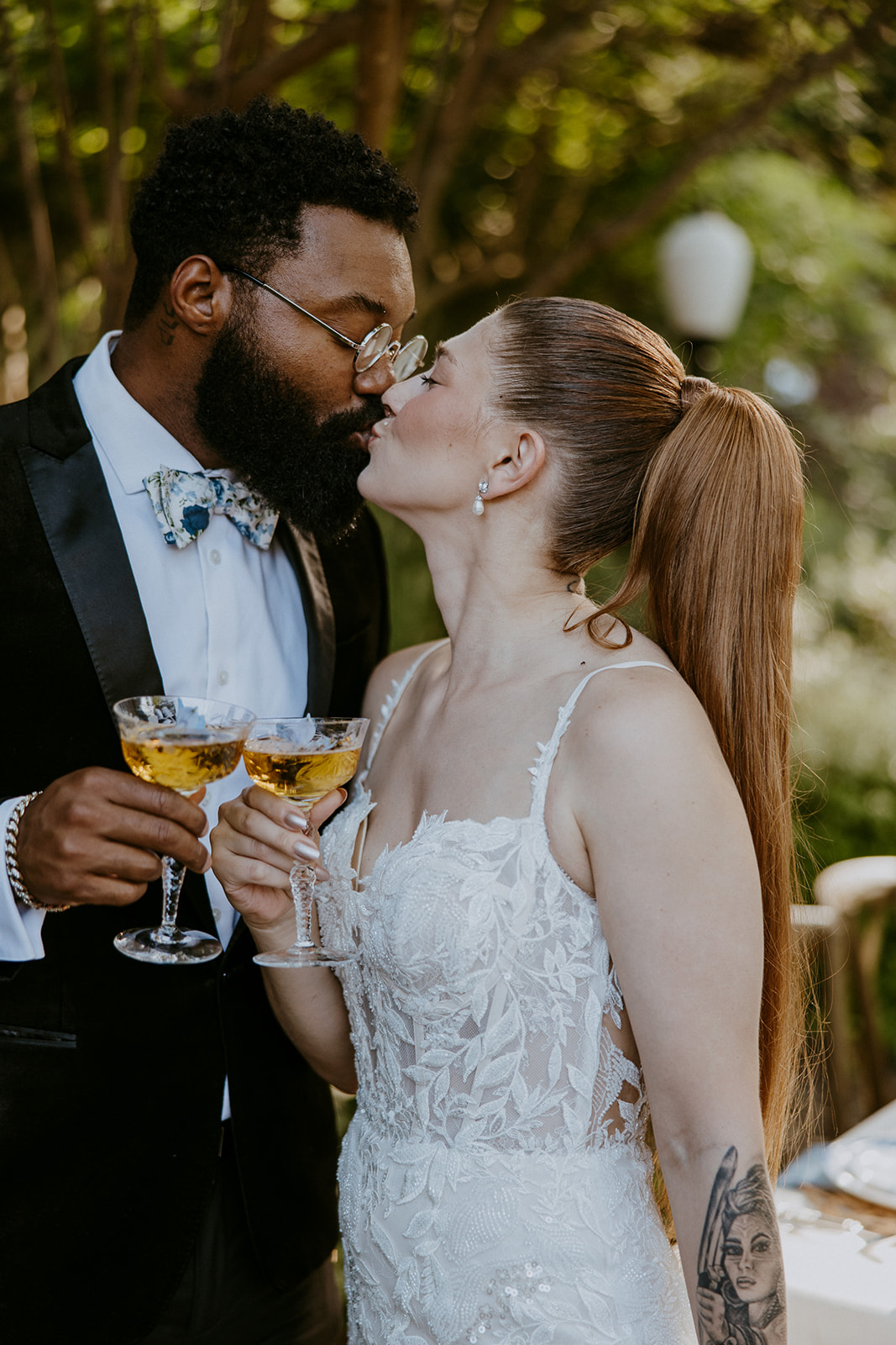 A bride and groom share a tender moment outdoors, with the bride holding a bouquet and the groom touching her chin. A table with floral decorations and candles is in the background.