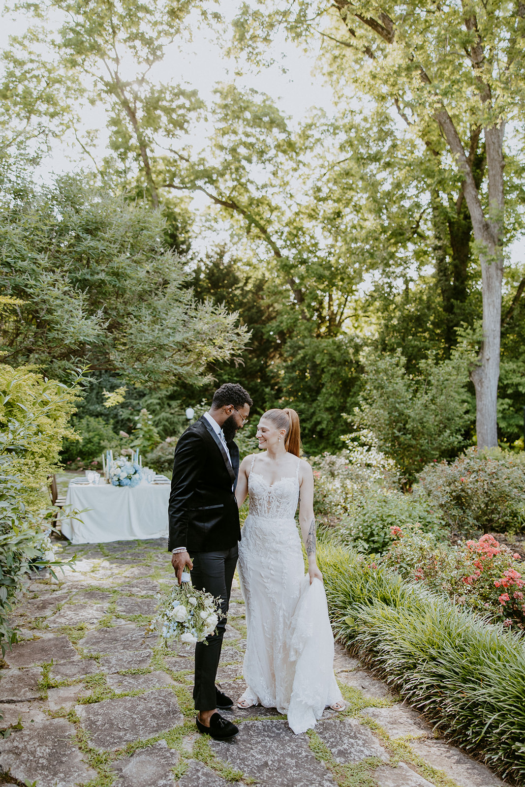 A couple in wedding attire stands on a stone path in a lush garden, holding hands and looking at each other.