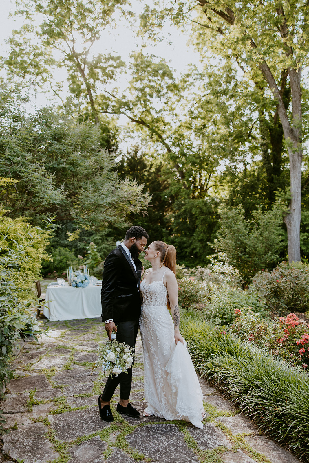 A couple in wedding attire stands on a stone path in a lush garden, holding hands and looking at each other.